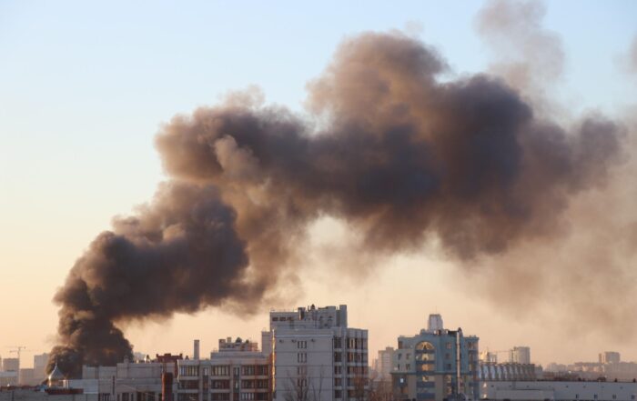 Factory is on fire , a number of white buildings surround it in an industrial park. Large black smoke clouds fill the air on a blue sky, sunshine day